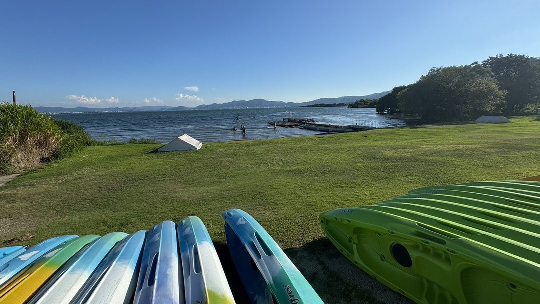 琵琶湖沿いの芝生の原っぱ。手前にカヌーが並び、向こうに湖と山並みが広がる